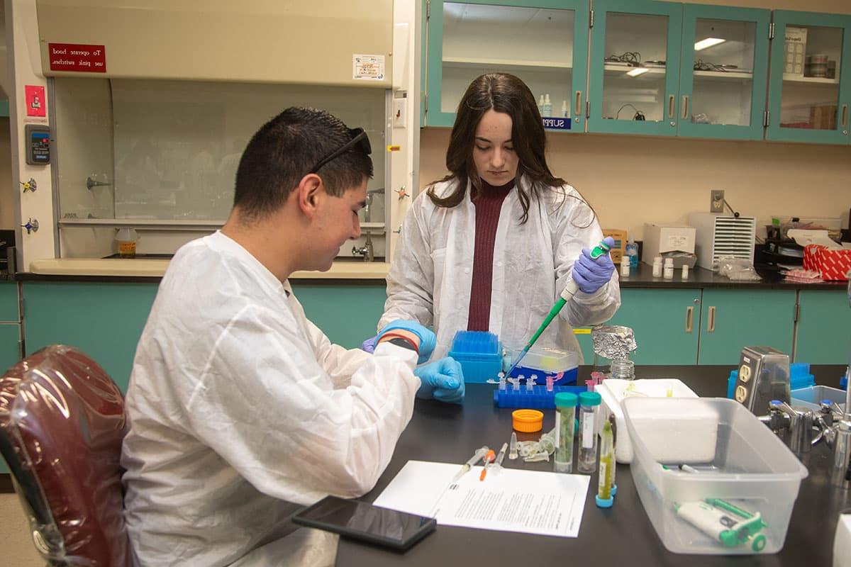 Two students in lab coats work with pipettes and test tubes at a table in a lab classroom